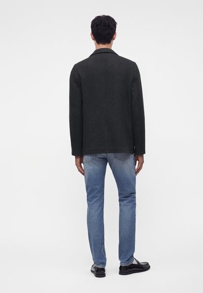 Homme debout, dos à la caméra, portant un manteau gris foncé, un jean bleu clair et des chaussures noires sur un fond blanc uni.