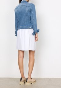 Vaaleansininen farkkutakki, jossa on sovitettu muotoilu, pitkät hihat ja nappikoristeet. Käytetty valkoisen mekon päällä, yhdistettynä beigenvärisiin slip-on-kengiin.