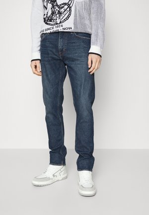 Donkerblauwe denim jeans met een rechte fit, met zichtbare stiksels en een klassiek vijf-pocket ontwerp, gecombineerd met witte sneakers.