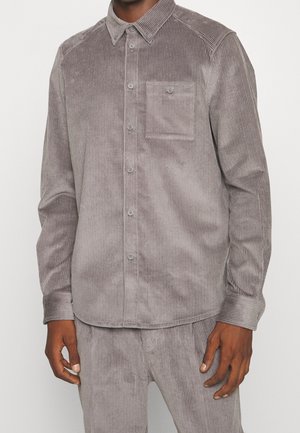 Man draagt een lichtbruin corduroy overhemd met lange mouwen, knopen, een borstzak en bijpassende corduroy broek.