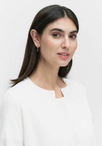 Witte top met een ronde halslijn en een gestructureerde afwerking. Het model heeft lang, donker haar en draagt kleine ringoorbellen. De achtergrond is vlak.