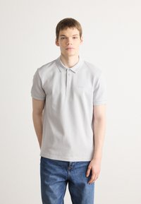 Polo shirt gris clair à manches courtes, avec des rayures texturées, un rabat à trois boutons et un logo discret sur la poitrine. Porté avec un jean en denim bleu.