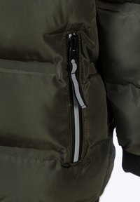 Olivgrön pufferjacka med en ficka med dragkedja som har kontrasterande vit och svart dragkedjeskon, strukturerad nylonväv och quiltad design.