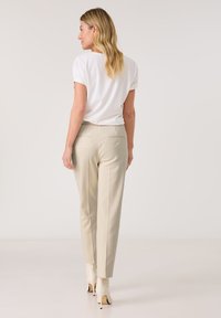 Beige maßgeschneiderte Hose mit glatter Textur, ausgestattet mit zwei Gesäßtaschen und Knopfakzenten. Kombiniert mit einem weißen kurzärmeligen Hemd.