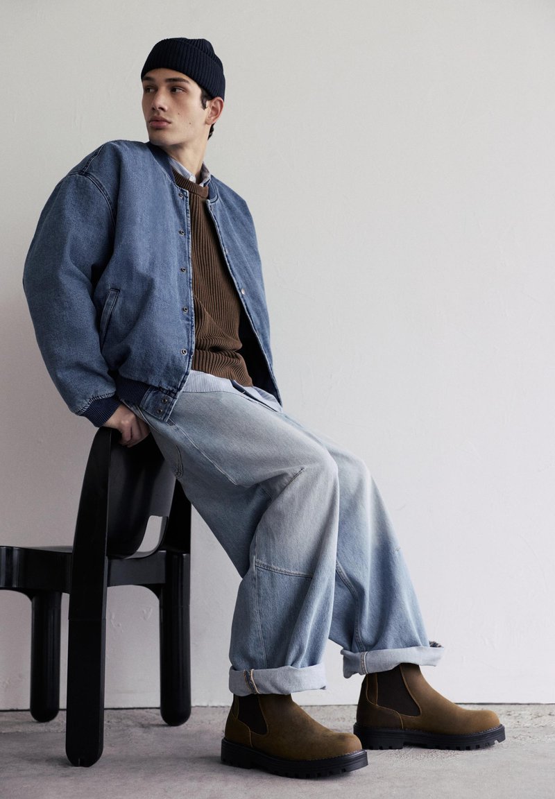Jeune homme portant un bonnet foncé, une veste en jean, un pull marron, un jean clair ample et des bottes marron, assis sur une chaise noire contre un mur blanc.