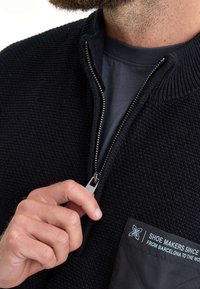 Schwarzer, strukturiert gestrickter Pullover mit Reißverschlussfront, ausgestattet mit einer Pattentasche und weißem Text. Der Reißverschluss-Zipper ist silberfarben und setzt einen Akzent im Design.