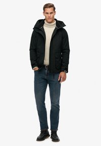 Schwarze Kapuzenjacke über einem cremefarbenen Strukturstrickpullover, kombiniert mit blauen Jeans und schwarzen Schnürschuhen. Verfügt über zwei Fronttaschen.
