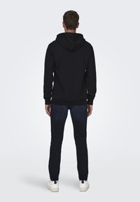 Hanorac negru cu glugă, mâneci lungi și mansete din material elastic, asociat cu blugi slim-fit de culoare bleumarin și adidași albi, văzut din spate.