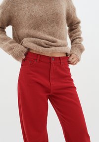 Jeans de mezclilla rojos con un corte recto y cinco bolsillos, combinados con un suéter de lana suave de color marrón claro. Los jeans cuentan con un cierre de botón.
