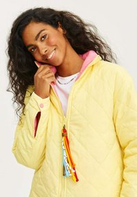 Žlutá prošívaná bunda s růžovým límcem, se zapínáním na zip vpředu a barevnými třásněmi. Texturovaná tkanina s volným střihem.