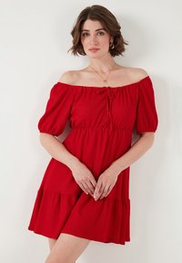 Red off-the-shoulder dress with puff sleeves, cinched waist, and tiered hem. Made of textured fabric, featuring a front tie detail.