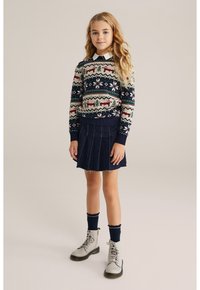 Stickad tröja med festliga mönster i grönt, rött och marinblått, kombinerad med en blå denimplisserad kjol och grå snörskor.