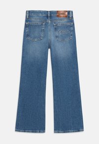 Jeans de ganga azul com corte flare e cintura média. Apresenta dois bolsos traseiros e um rótulo de cintura em couro marrom texturizado. Detalhe de costura clássica.
