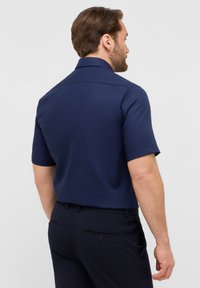 Camicetta blu navy a maniche corte con colletto classico, tessuto liscio, dettaglio della cucitura del retro e vestibilità comoda. Abbinata a pantaloni scuri.