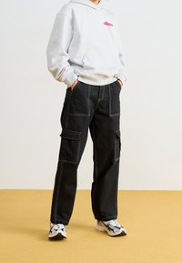 Persona che indossa un hoodie grigio chiaro di ellesse, pantaloni cargo neri con cuciture bianche e sneakers bianche e nere, in piedi su un pavimento giallo.