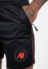 Shorts noirs en tissu lisse, avec une bande latérale rouge et un logo circulaire affichant un graphique de gorille avec du texte rouge et noir.