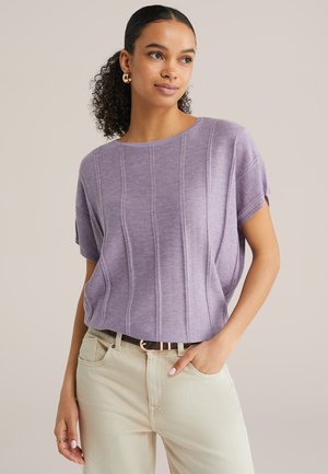 Kobieta o kręconych włosach, nosząca złote kolczyki-koła, liliowy, teksturowany sweter z krótkim rękawem oraz jasnobeżowe spodnie z czarnym paskiem.