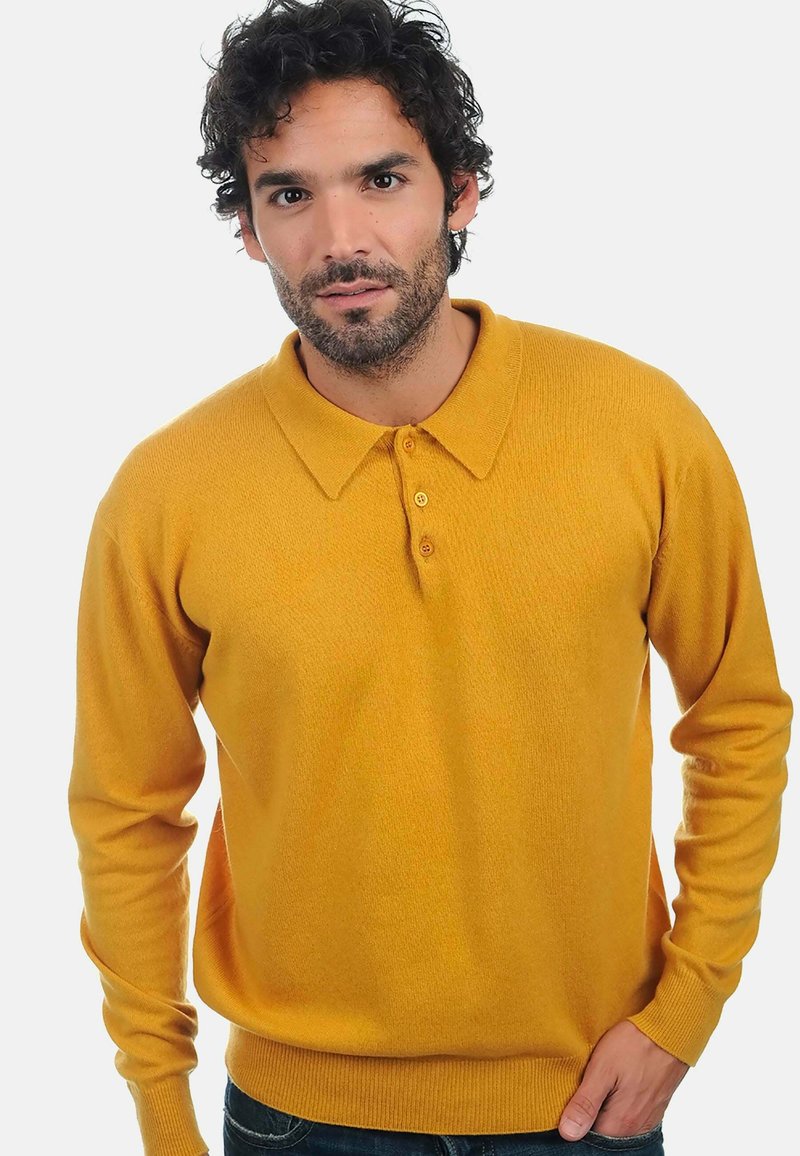 Mann mit dunklem, lockigem Haar und Bart, der einen senfgelben Langarm-Polo-Pullover mit Knöpfen trägt und mit einer Hand in der Tasche steht.