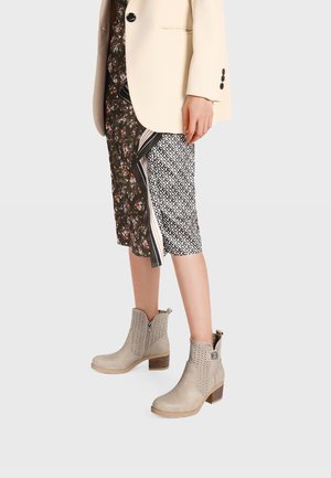 Donna con stivaletti beige con tacco, gonna midi con motivi floreali e geometrici, e blazer di colore chiaro.