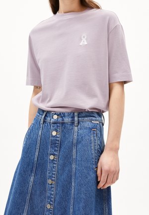 Frau trägt ein fliederfarbenes kurzärmeliges T-Shirt mit kleinem weißem Logo und einen hoch taillierten Jeansrock mit Knöpfen vorne und Taschen.