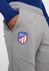 Pantalones deportivos grises con un logo bordado que presenta rayas rojas y blancas y acentos azules. Se pueden ver bolsillos laterales y una cintura con cordón.