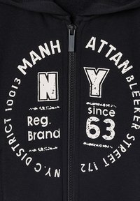 Schwarze Kapuzenjacke mit Reißverschluss, versehen mit weißen Texten und Grafiken, die Städtenamen, Zahlen und einem runden Logodesign zeigen. Das Material wirkt weich und strukturiert.