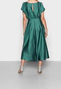 Robe en satin verte avec un dos à découpe, manches courtes, taille cintrée et jupe fluide. Des escarpins argentés à brides de cheville complètent le look.