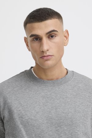 Noor mees lühikeste tumedate juustega, kandes heledat halli ümar-kaelusega pusa, taustal hele üksildane sein.