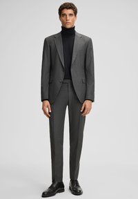 Uomo che indossa un abito grigio a righe sottili con un dolcevita nero e scarpe eleganti nere, in piedi contro uno sfondo grigio chiaro uniforme.