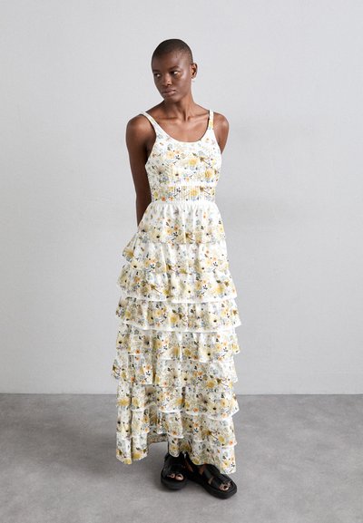 Robe maxi fleurie avec un fond blanc, des fleurs jaunes et bleues, un corsage froncé et des volants à plusieurs niveaux. Des sandales noires complètent le look.