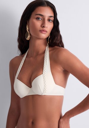 Femme portant un haut de bikini blanc clouté à col halter et de grandes boucles d'oreilles en or, avec de longs cheveux foncés et une expression neutre sur fond uni.