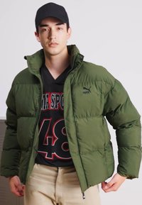 Giacca a vento verde con colletto alto, caratterizzata da sezioni trapuntate. Abbinata a una maglietta nera con grafica e pantaloni beige a vestibilità standard.