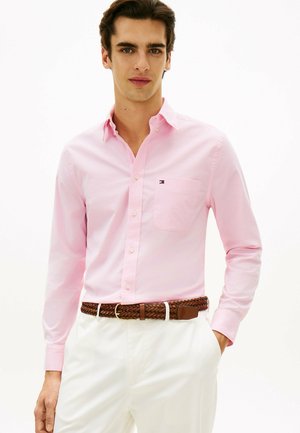 Młody mężczyzna w jasnoróżowej koszuli na guziki i białych spodniach z brązowym plecionym paskiem, stojący z jedną ręką w kieszeni.