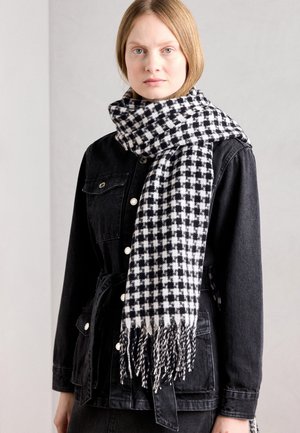 Écharpe à carreaux noir et blanc en tissu doux, avec des bords frangés. Portée avec une veste en jean noire, ajoutant texture et contraste.