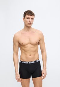 Άνδρας με κοντά καστανά μαλλιά φοράει μαύρα μπόξερ Puma, στέκεται μπροστά σε απλό ανοιχτό γκρι φόντο, κοιτάζοντας μπροστά με ουδέτερη έκφραση.