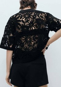 Black crochet top featuring floral patterns, short sleeves, and an open back design, paired with black shorts. Textured, lace-like appearance.