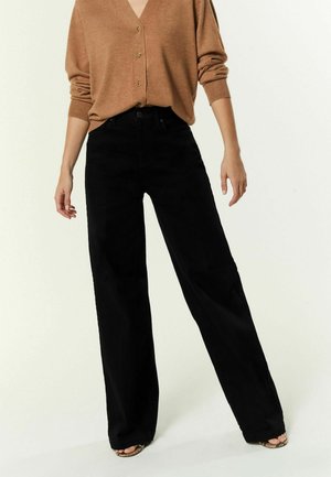 Femme portant un pantalon noir ample, un cardigan beige boutonné et des chaussures ouvertes, debout sur un fond clair.
