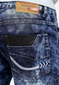 Jeans in denim con una sfumatura di blu sbiadito, caratterizzati da una toppa in pelle nera sulla tasca posteriore e cuciture bianche a forma di curve e frecce.
