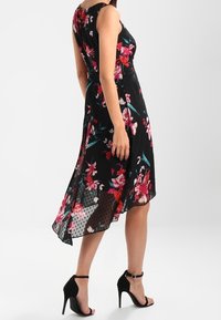 Czarna sukienka w kwiaty z asymetrycznym dołem, kwiatowym nadrukiem w czerwonym i turkusowym kolorze oraz lekką, przezroczystą tkaniną. Połączona z czarnymi szpilkami.