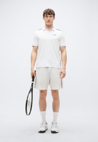 Weißes sportliches Polohemd mit schwarzen Akzenten, kombiniert mit passenden Shorts. Das Modell hält einen Tennisschläger und trägt weiße Schuhe und Socken.
