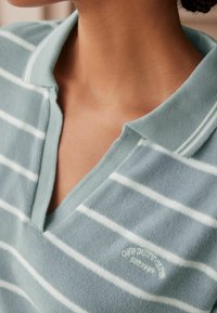 Helles blaues, kurzärmliges Poloshirt mit weißen horizontalen Streifen. Hat einen V-Ausschnitt, eine weiche Textur und bestickte Aufschrift "Off Duty Club".