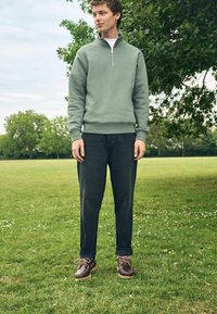 Sudadera verde con cremallera, jeans oscuros, zapatos marrones con cordones. Entorno al aire libre con hierba y árboles de fondo. Cielo ligeramente nublado.