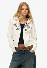 Sudadera con cremallera color marfil con letras "Superdry", bolsillos frontales y capucha con cordón, combinada con una camiseta negra y jeans de denim azul holgados.