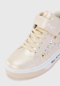 Crème-kleurige sneaker met klittenbandsluiting, veters, metallic studs en een witte zool met decoratieve tekst op de zijkant.