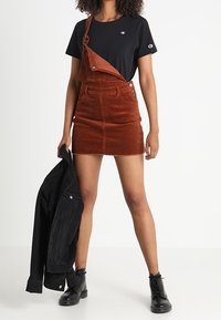 T-shirt noir, robe salopette en velours côtelé marron avec poches et boutons, associée à une veste en jean noire et des bottines noires.