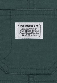 Dunkelgrüner Stoff mit strukturierter Oberfläche, auf dem ein weißes Etikett genäht ist, das "LEVI STRAUSS & CO." sowie Produktdetails zeigt.