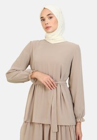 Beige, långärmad klänning med samlade ärmar och knytmidja. Tillverkad av lätt tyg, med en lagerdesign och en mjuk textur.