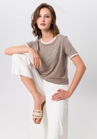 Lichtbruine t-shirt met korte mouwen en witte bies, gecombineerd met witte wijde broeken en beige instappers met ronde accenten.