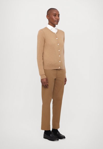 Beige knit cardigan with round neckline, button front, and long sleeves paired with brown tailored trousers. Shoes are black loafers.