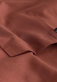 Tissu marron à texture lisse, avec des bords pliés. Un côté présente une poche et des détails de couture. Motif de tissage subtil visible.